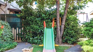 Un parque infantil con un tobogán verde y asideros naranjas, rodeado de exuberante vegetación y una jardinera de piedra.