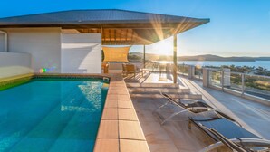 Un área de piscina en la azotea con vistas al océano y colinas, rodeada de sillones de descanso y un espacio de estar cubierto.