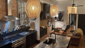 A modern kitchen with a gas stove, dark cabinetry, and a large island. There is a dining area with a marble table and a sofa. The room features exposed brick walls and wooden beams. - Luxurious industrial-chic loft in historic downtown Sheridan. This stunning 2-bedroom, 2-bath apartment sleeps up to 9, featuring 2 king suites, 2 1/2 bathrooms sleeping alcove, modern kitchen, fireplace, & Main Street balcony views. (Sheridan)