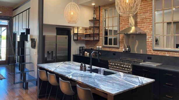 A modern kitchen with a marble island, stainless steel appliances, and a brick accent wall. - Luxurious industrial-chic loft in historic downtown Sheridan. This stunning 2-bedroom, 2-bath apartment sleeps up to 9, featuring 2 king suites, 2 1/2 bathrooms sleeping alcove, modern kitchen, fireplace, & Main Street balcony views. (Sheridan)