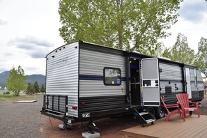 Een grijze en zwarte camper met een schuifdeur, een klein aangebouwd gedeelte en een rode stoel op een houten terras.