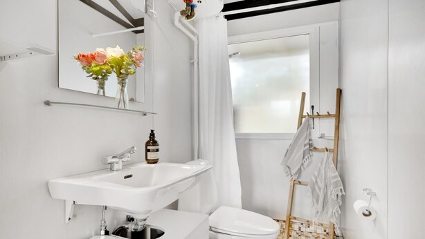 Une salle de bain moderne avec un lavabo blanc, un miroir et des toilettes. On y trouve un rideau de douche, un porte-serviettes en bois et une fenêtre qui laisse entrer la lumière naturelle.
