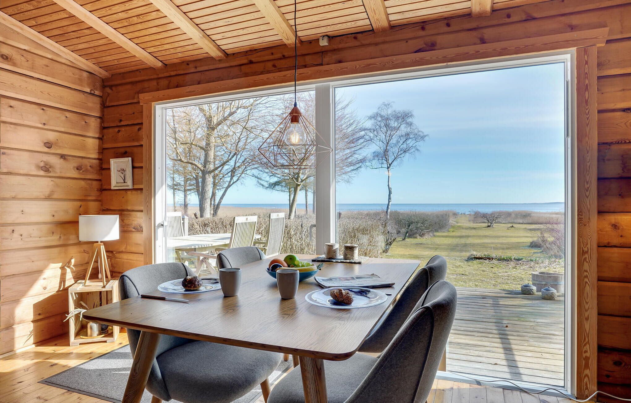 A wooden cabin with a dining table and chairs, a lamp, and a view of the beach and trees.