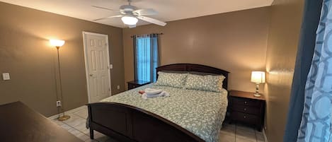 Une chambre à coucher avec un lit, un ventilateur de plafond, une table de chevet, une lampe et une fenêtre avec des rideaux.