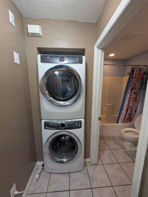 A compact laundry room with two stacked front-loading washing machines, a shower area with a floral curtain, and a toilet.