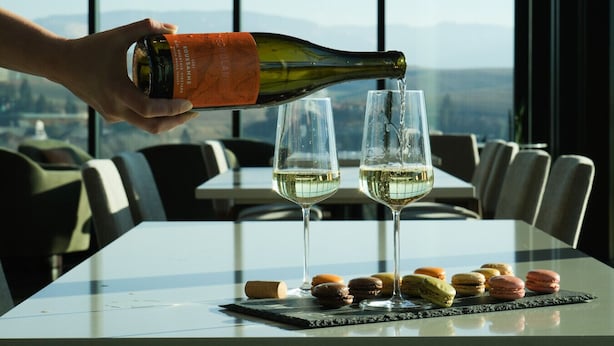 A hand pouring wine into two glasses on a table with macarons, in a room with large windows and a view of the city.