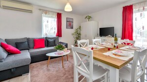 Un salon moderne avec un canapé gris, des coussins rouges, une petite table ronde ornée d’une plante, et un coin repas avec des chaises blanches et une table en bois dressée pour deux personnes.