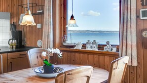 Un intérieur en bois offrant une vue sur la mer, une table à manger avec des chaises et un coin cuisine équipé d’une machine à café et d’ustensiles.