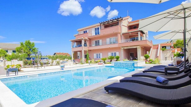 A swimming pool with lounge chairs and umbrellas, surrounded by a pink building with balconies. - Gorgeous apartment in Grebastica with WiFi (Grebastica)