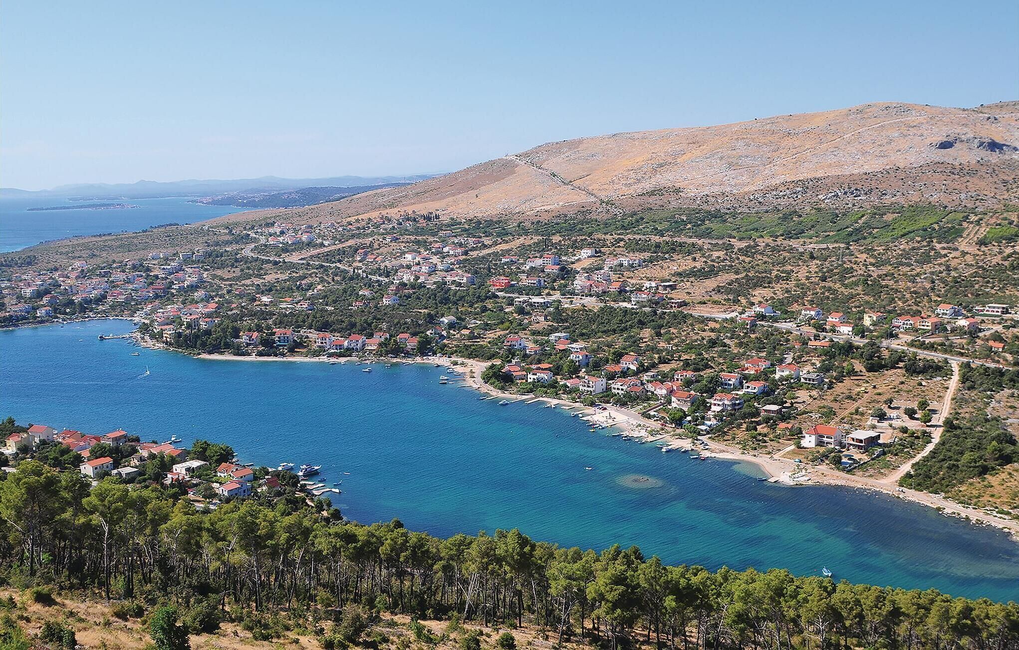 A coastal town with houses along the shoreline, a clear blue sea, and a hilly backdrop.