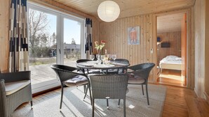 Una habitación acogedora con una mesa redonda dispuesta para comer, sillas de mimbre, un ventanal con cortinas y una cama en la habitación contigua.