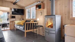 Interior acogedor de una cabaña con un sofá, un televisor montado en la pared, un estufa de leña y un ventanal con vistas a árboles.