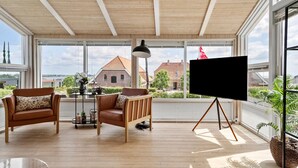 Une pièce spacieuse avec de grandes fenêtres, un plafond en bois, deux fauteuils en cuir, un lampadaire et un téléviseur posé sur un trépied.