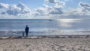 Une plage où l’on voit une personne et un enfant se promener le long du rivage, un bateau au loin et un ciel dégagé avec quelques nuages.