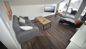 A modern living room with a grey sofa, a wooden coffee table, and a black armchair. A flat-screen TV is mounted on the wall, and the room has large windows offering a view of the outside. - Familienferienhaus Mare Frisium, Meerblick, am Deich, Waschtrockner, Sauna, Ofen (Wangerland)