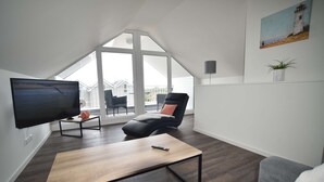A modern living room with a flat-screen TV, a wooden coffee table, a black leather chair, and a grey sofa. - Familienferienhaus Mare Frisium, Meerblick, am Deich, Waschtrockner, Sauna, Ofen (Wangerland)