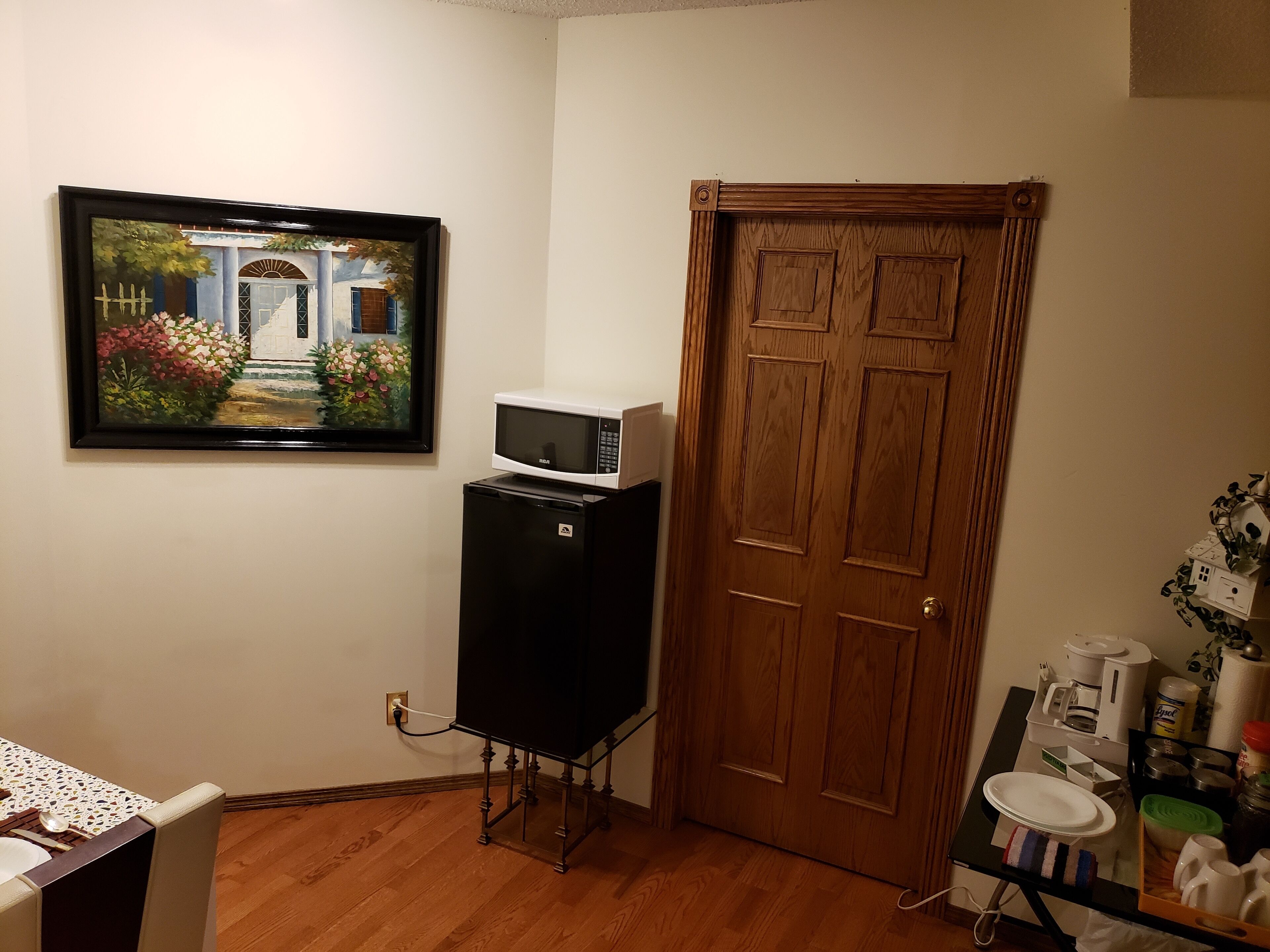 Ein Zimmer mit Holztür, einem schwarzen Kühlschrank mit integriertem Mikrowellenherd, einem kleinen Tisch mit Teller und Tassen sowie einem gerahmten Bild an der Wand.