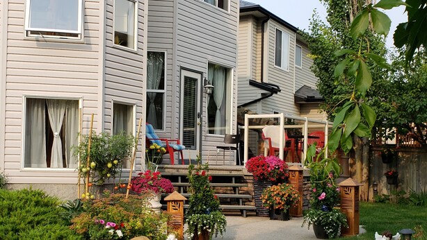 A two-story house with a front porch, a small garden with various plants and flowers, and a wooden deck. - PRIVATE 1 B/R Suite in Upscale formerShowhome  . 4 MIN walk to Lakeside beach Pk (Chestermere)
