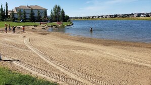 A sandy beach with a lake, a playground, and houses in the background. - PRIVATE 1 B/R Suite in Upscale formerShowhome  . 4 MIN walk to Lakeside beach Pk (Chestermere)