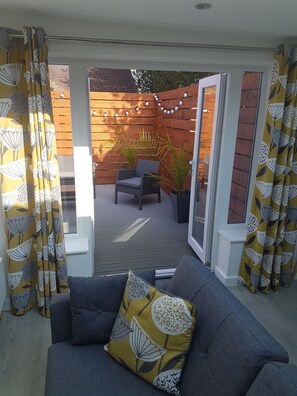 A modern living room with a grey sofa, a yellow patterned throw pillow, and a wooden deck area with a chair and plants. - The Coneygree @ Northwick (Worcester)