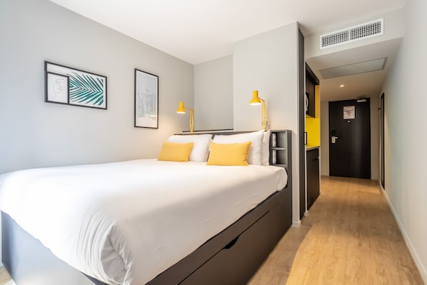 Una habitación de hotel moderna con cama, lámparas de noche, un pequeño escritorio y una puerta que da a un pasillo.