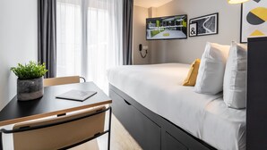 A modern hotel room with a bed, a desk, a chair, a potted plant, a TV on the wall, and a window with curtains.