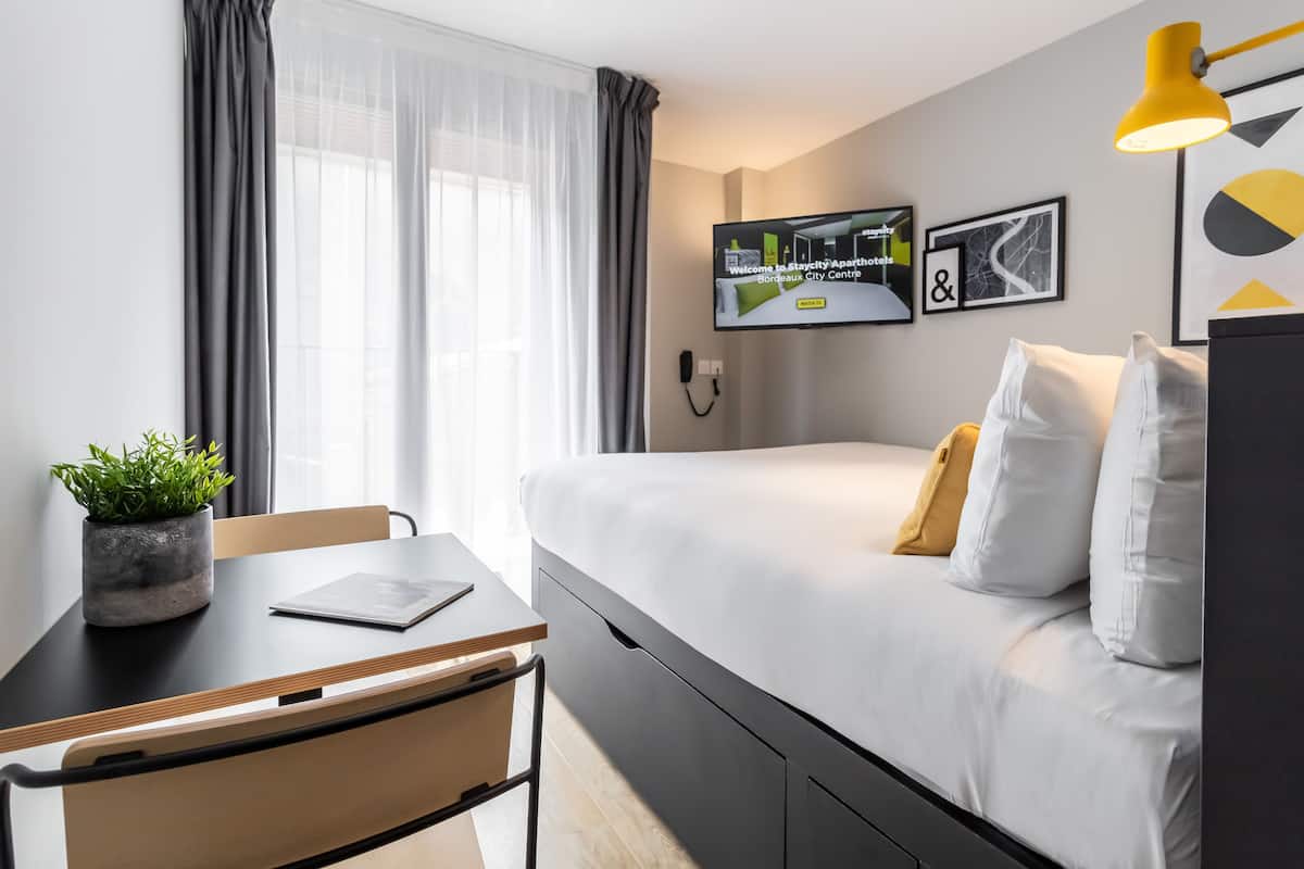 Ein modernes Hotelzimmer mit Bett, Schreibtisch, Stuhl, einer Topfpflanze, einem an der Wand montierten Fernseher und einem Vorhangfenster.
