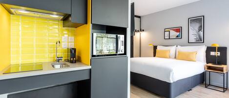 Habitación compacta con kitchenette, una cama con dos almohadas y una mesita de noche con lámpara.