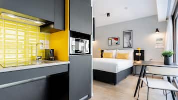 Habitación de hotel compacta con kitchenette, cama con ropa blanca, una pequeña mesa de comedor y una planta en maceta.