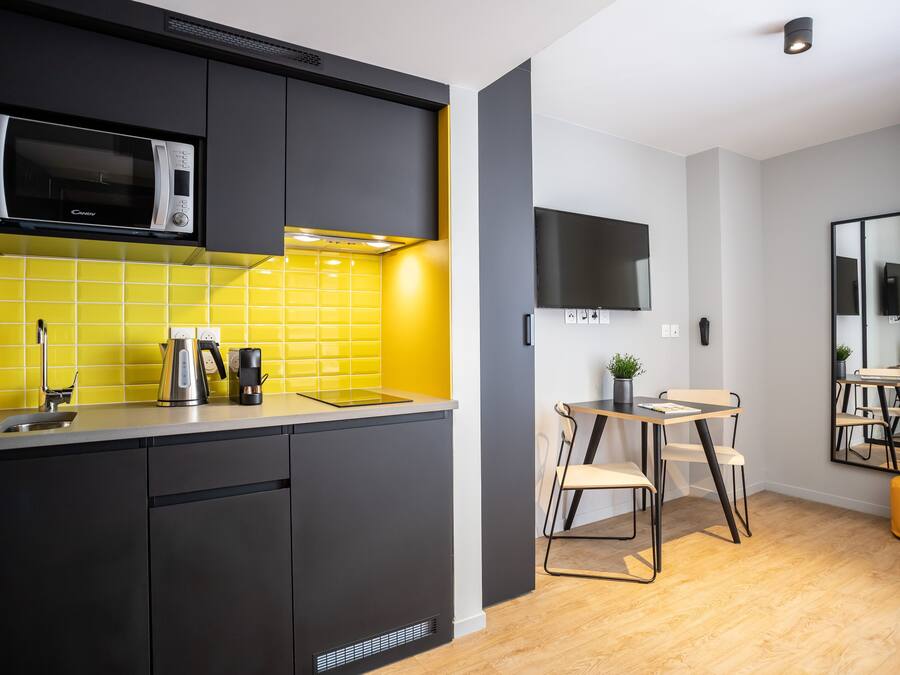 A modern kitchen with a microwave, kettle, and coffee maker on the counter, a dining area with a table and chairs, and a flat-screen TV mounted on the wall.