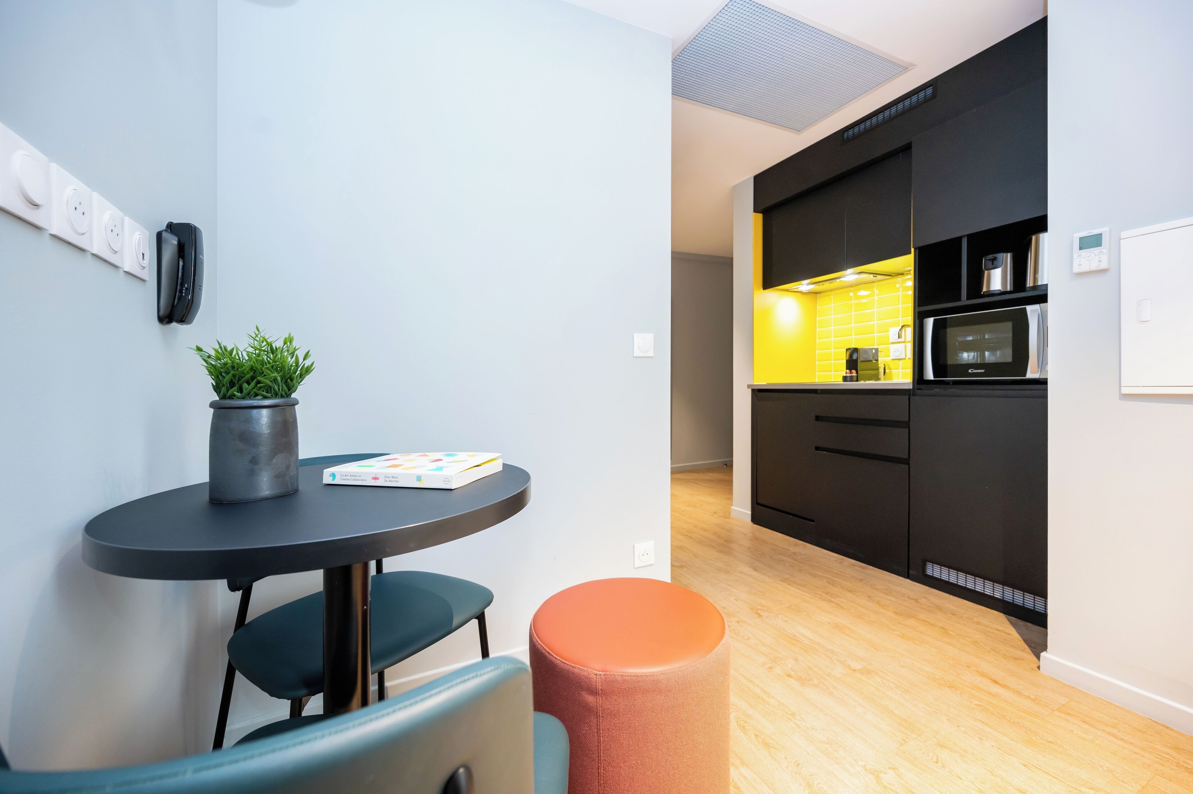 A modern kitchen with black cabinets, a microwave, and a coffee machine. There is a small dining area with a table and chairs, and a potted plant on the table.