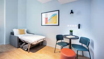 Une pièce de taille modeste comprenant un lit, une petite table avec des chaises, un luminaire fixé au mur et une prise de courant.