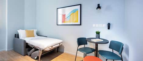 A compact room with a bed, a small table with chairs, a wall-mounted light, and a power outlet.