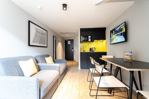 Una sala de estar moderna con un sofá, una mesa de comedor con sillas, una zona de cocina con microondas y un televisor montado en la pared.