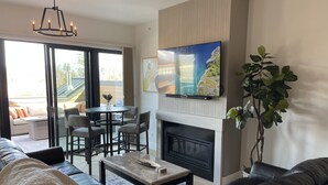 A modern living room with a fireplace, a flat-screen TV mounted above it, and a dining area with a round table and chairs.