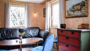 Ett rum med en väggpanel av trä, en svart lädersoffa, en blå stol, ett träbord, en röd och grön byrå och en målning som föreställer ett hus vid en sjö.