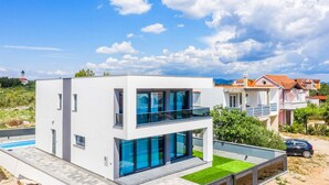 A modern two-story house with large glass windows and a balcony, surrounded by a well-maintained lawn and a clear blue sky.