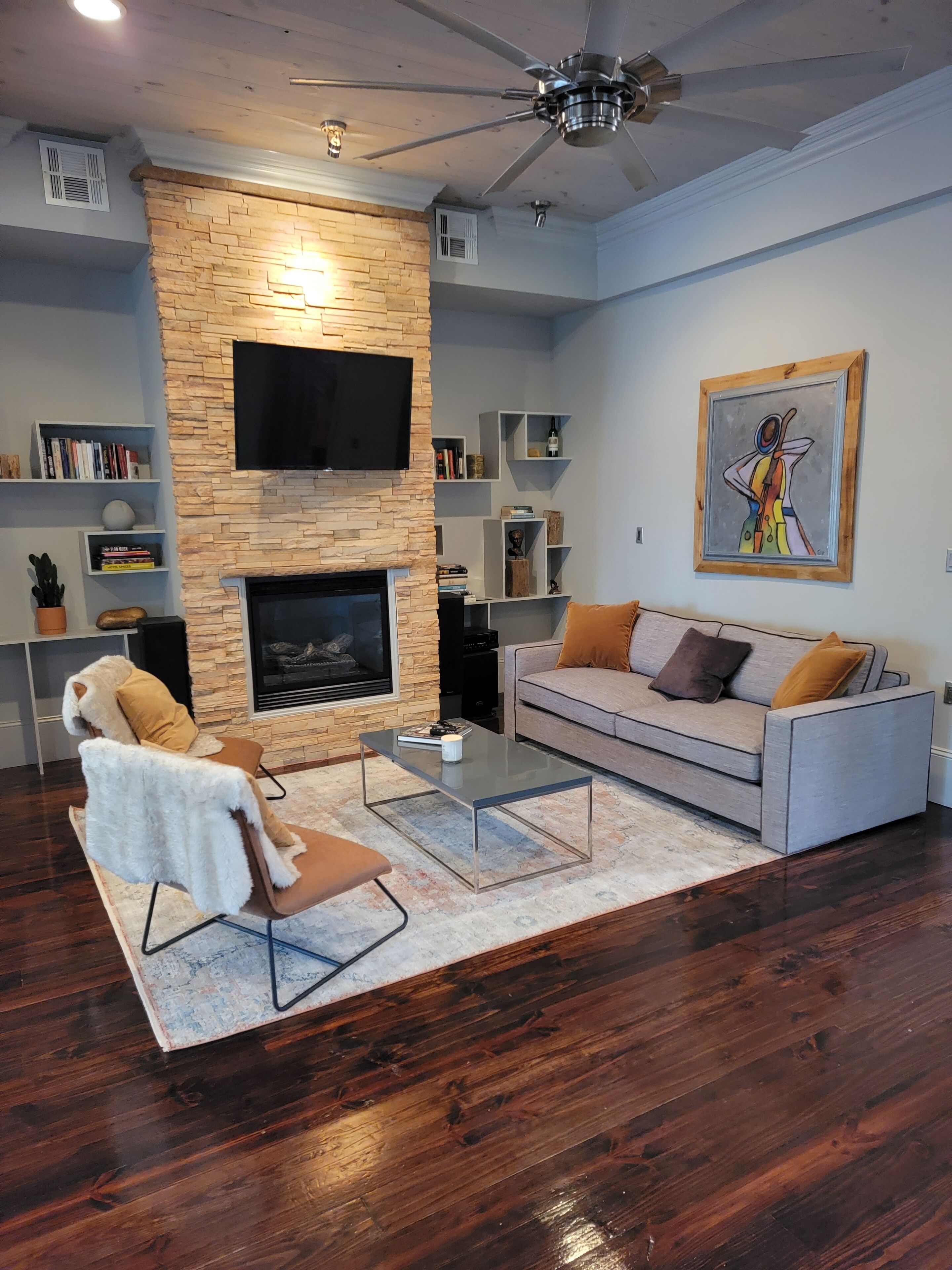 A living room with a stone fireplace, a flat-screen TV mounted above it, a grey sofa with orange cushions, a glass coffee table, and a ceiling fan.
