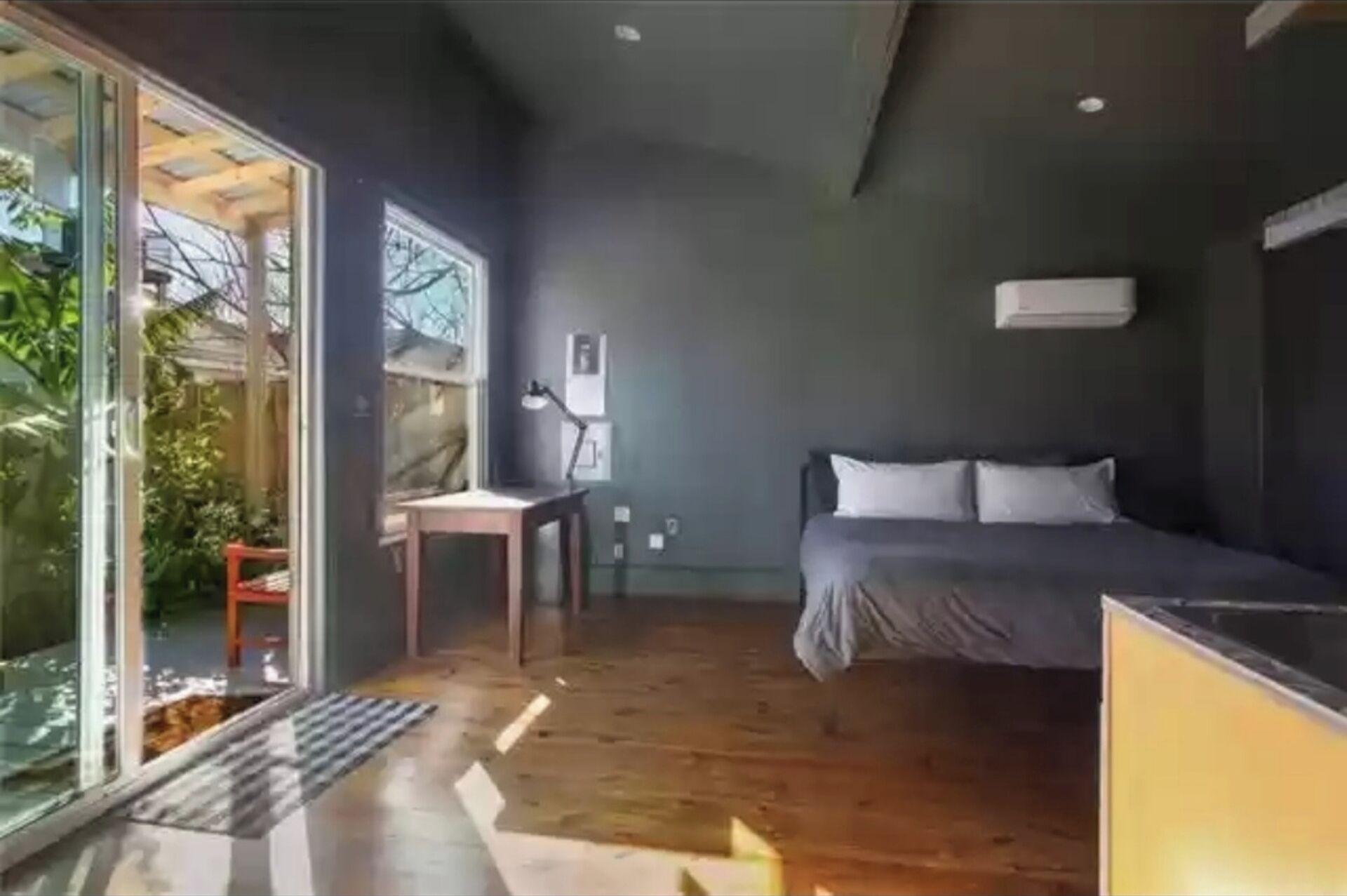 Un dormitorio moderno con cama, escritorio y silla. Hay una ventana que proporciona luz natural y vistas a la vegetación exterior.