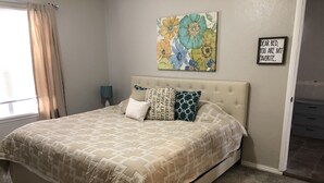 A bedroom with a bed, a nightstand, a lamp, a floral artwork, and a framed message on the wall.