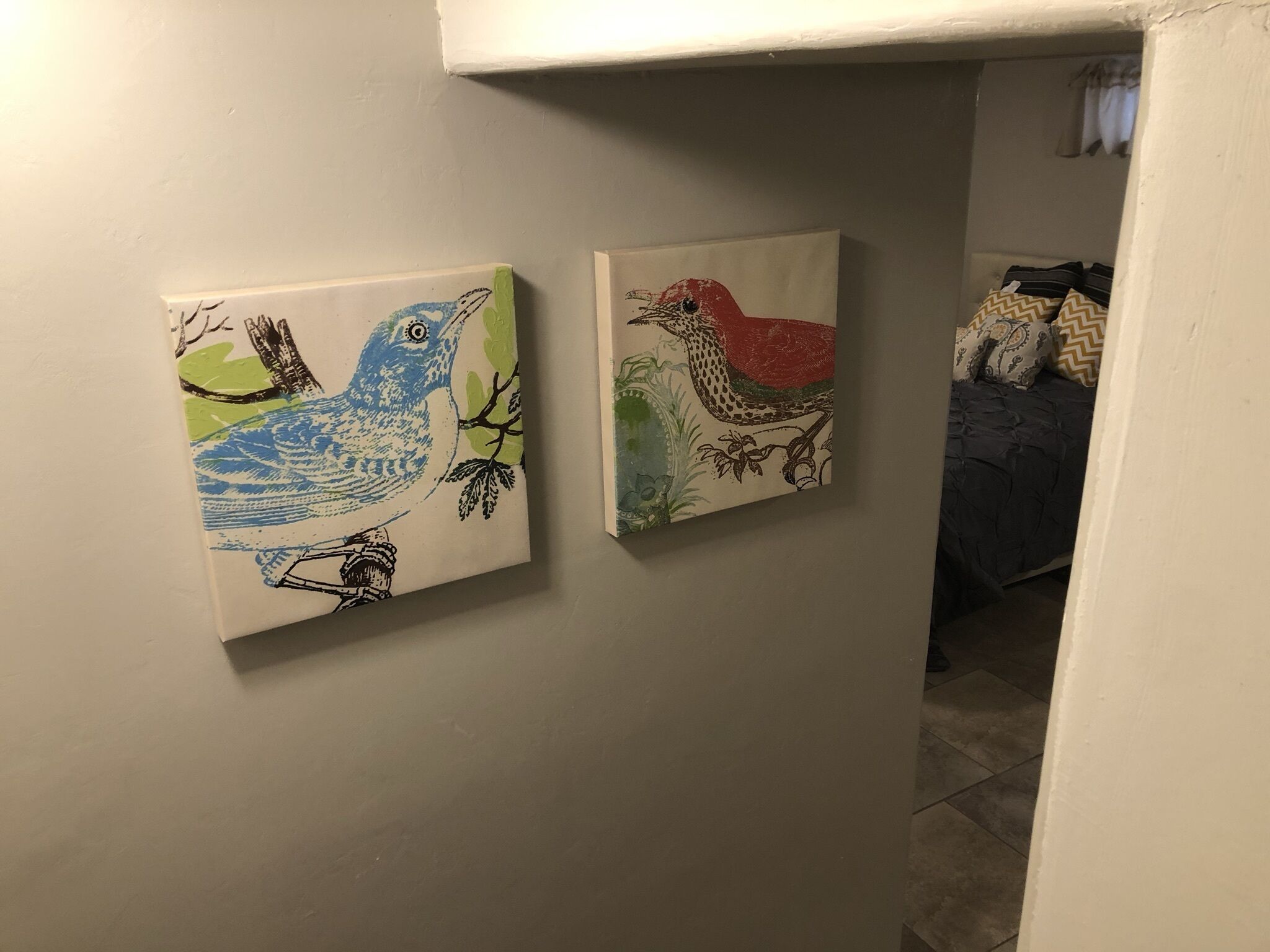 Deux tableaux représentant des oiseaux sont accrochés à un mur dans une pièce où se trouvent un lit et une fenêtre en arrière-plan.