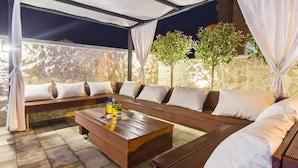 An outdoor seating area with a wooden bench and a table, surrounded by white cushions.