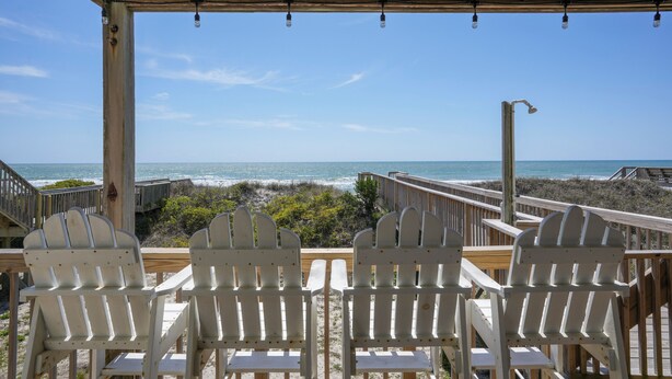 Una terraza de madera con sillas Adirondack blancas mirando a la playa.