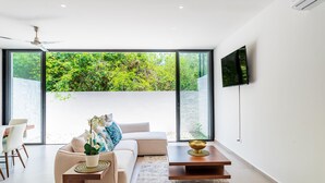 A modern living room with a large window, a flat-screen TV, a sofa, a coffee table, and a dining area with chairs. - Retreat into Exotic Paradise in Riviera Maya, 15 minute walk to beach (residencial El Cielo, Solaridad)