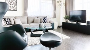 A modern living room with a sofa, a glass coffee table, a black leather chair, a flat-screen TV, and a floor lamp.
