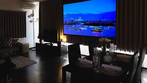 A modern living room with a large flat-screen TV mounted on the wall, a dark wood floor, a sofa, a coffee table, and a dining area with a table set for two.