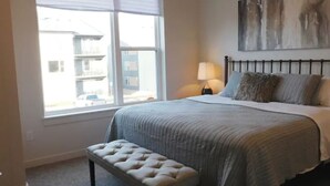 A bedroom with a large window, a bed with a grey bedspread, a bedside lamp, and a painting on the wall. - Brand new 2021 3 bedroom 2 bath condo. Great location just off interstate 90. (Livingston)