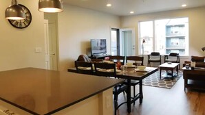 A modern kitchen with a central island, dining area with chairs, a TV, and a large window with a view of buildings. - Brand new 2021 3 bedroom 2 bath condo. Great location just off interstate 90. (Livingston)