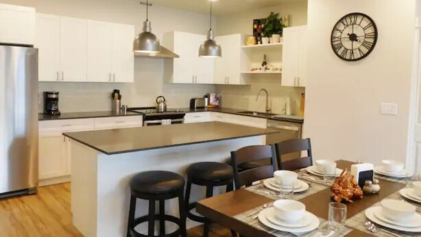 A modern kitchen with white cabinets, a central island with bar stools, pendant lighting, and a wall clock. - Brand new 2021 3 bedroom 2 bath condo. Great location just off interstate 90. (Livingston)