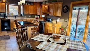 A modern kitchen with wooden cabinets, a dining area with a round table, and a view of a snowy landscape through a sliding glass door. - Farm Island Missouri River Views (Pierre)
